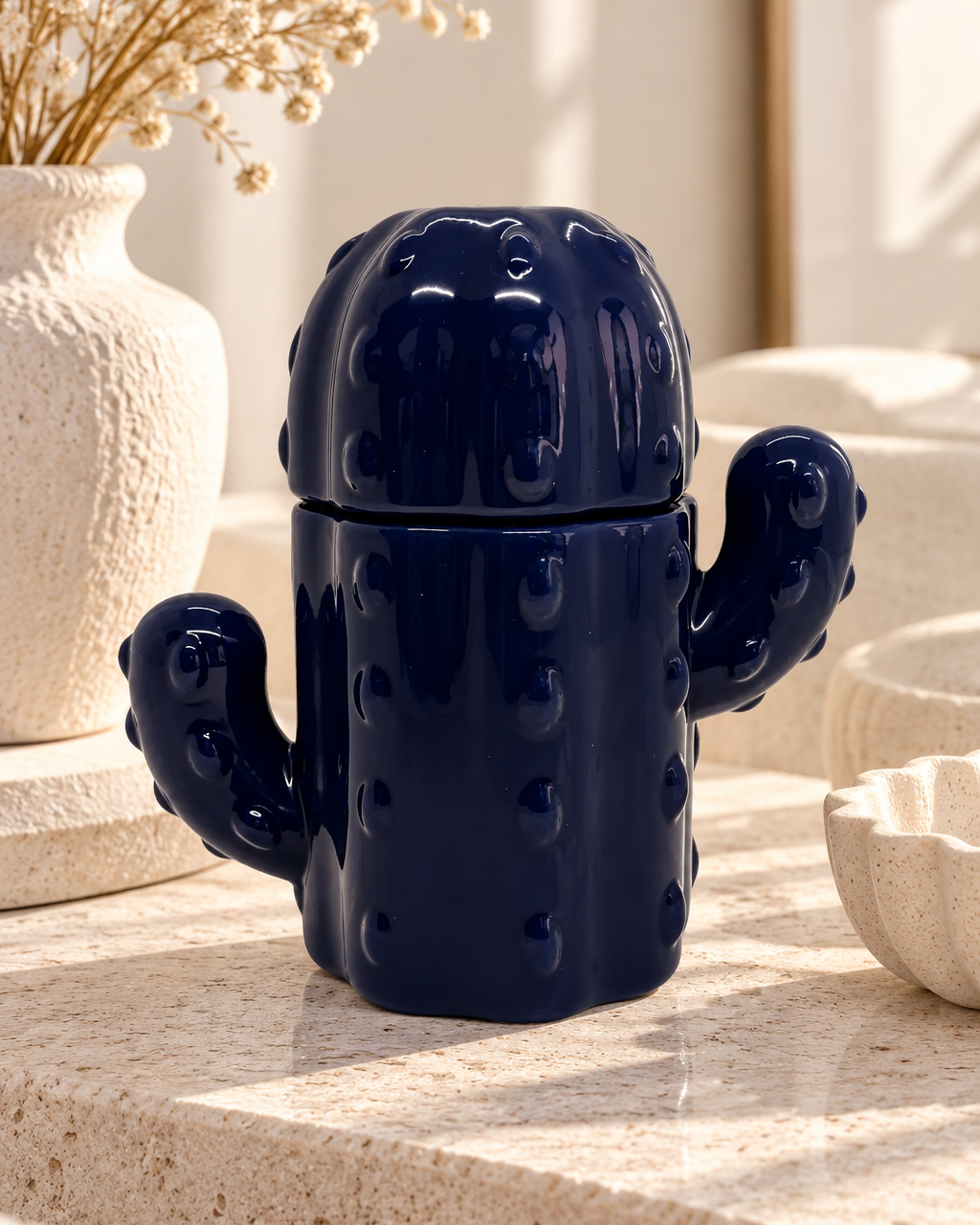 Boîte à allumettes en résine époxy bleu marine brillant, en forme de cactus avec deux bras et un couvercle arrondi, posée sur une surface en pierre beige dans un intérieur lumineux aux tons blancs et crème, près d'un vase et de fleurs séchées.