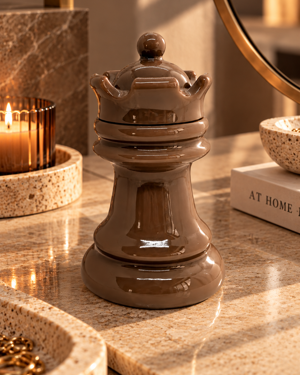 Boîte décorative en résine époxy taupe brillant, en forme de reine d’échecs couronnée, posée sur une console en pierre beige, près d’une bougie allumée et d’un miroir doré, dans une ambiance contemporaine chaleureuse.