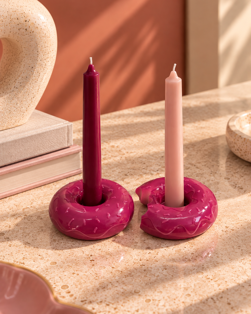 Duo de bougeoirs donuts en résine époxy rose fuchsia brillant, dont un croqué, accueillant deux bougies fines bordeaux et rose poudré, posés sur une table en pierre beige dans une ambiance design aux tons terracotta et pastel.