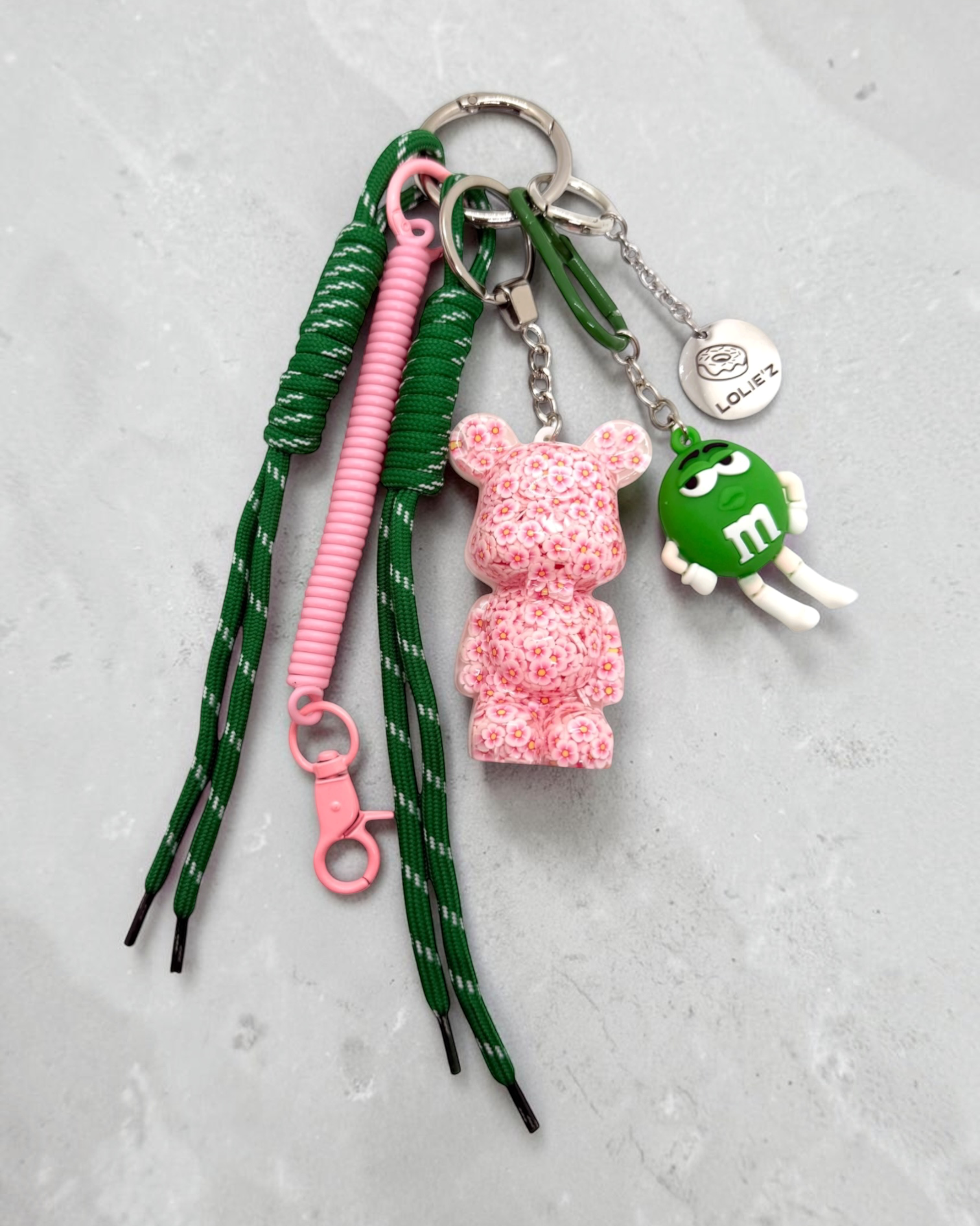 Grigri de sac Lolie’z rose et vert avec Teddy’z fleuri en résine époxy, cordons verts, mousqueton rose et breloque bonbon, assemblé à la main en France.