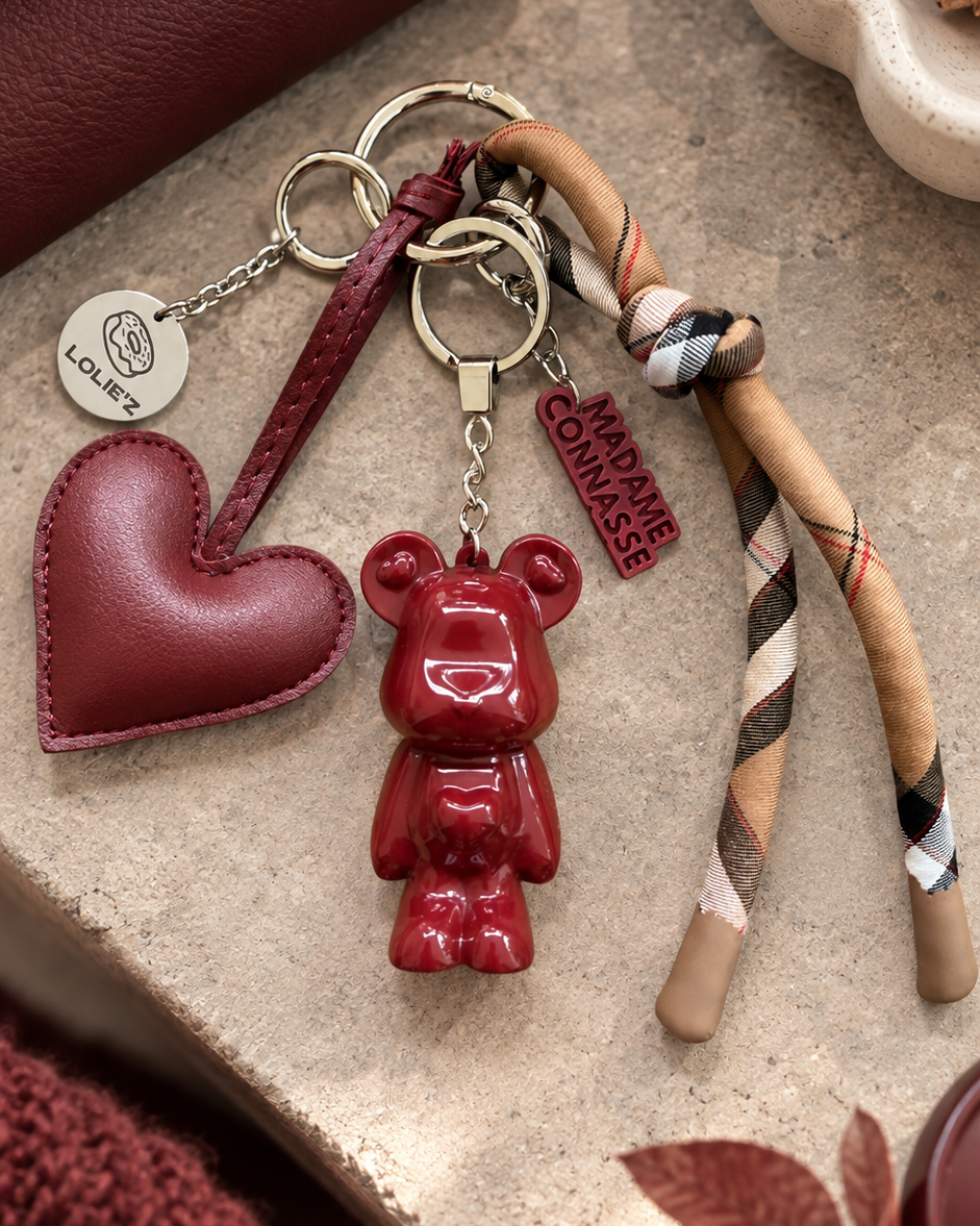 Grigri de sac Marley Onirique Lolie’z avec ourson rouge bordeaux en résine époxy, cœur, foulard à carreaux et médaille gravée, présenté dans une mise en scène chaleureuse.