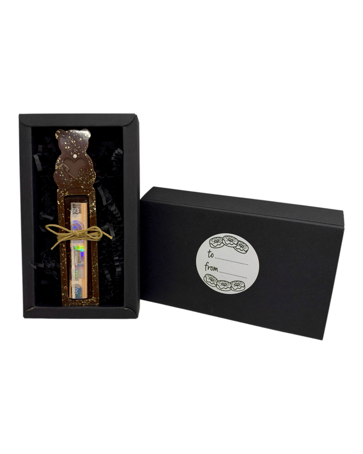 Support à billet de Noël Teddy Bear en forme d'ourson chocolat à paillettes dorées, avec billet roulé et nœud en ficelle, présenté dans son coffret noir avec étiquette cadeau.