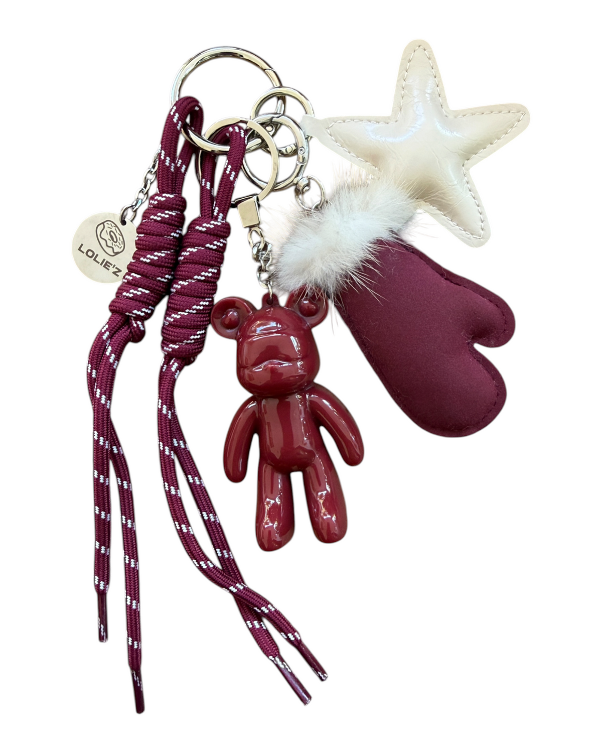 Grigri Urban Ted Miss Burgundy accroché à un sac à main taupe dans une ambiance hivernale cosy, avec ours bordeaux brillant, étoile écrue et moufle décorative.