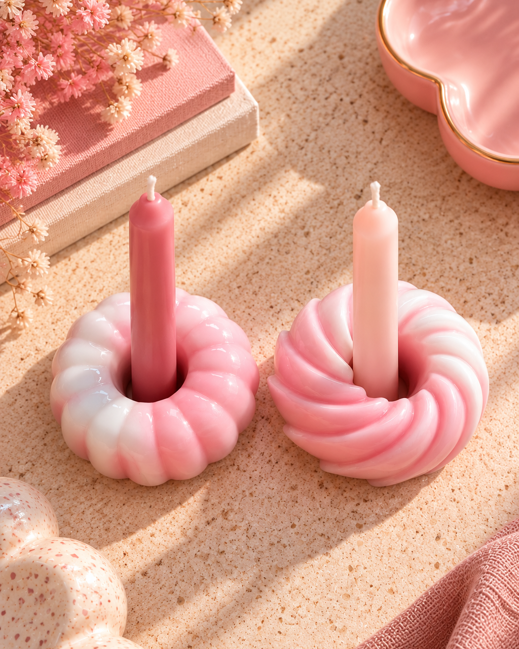 Deux bougeoirs en résine époxy rose et blanc, aux formes de donuts torsadés, accueillent des bougies roses sur une table beige dans une ambiance pastel moderne et lumineuse.