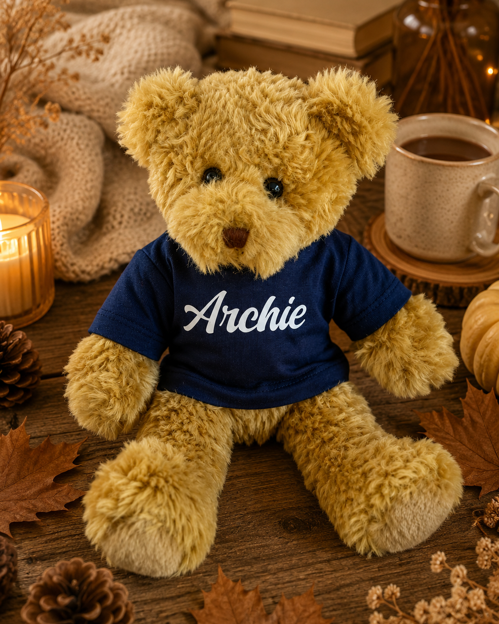 Ours en peluche Archie portant un t-shirt bleu marine personnalisé, photographié dans une ambiance cosy d’automne avec plaid, tasse et feuilles séchées.