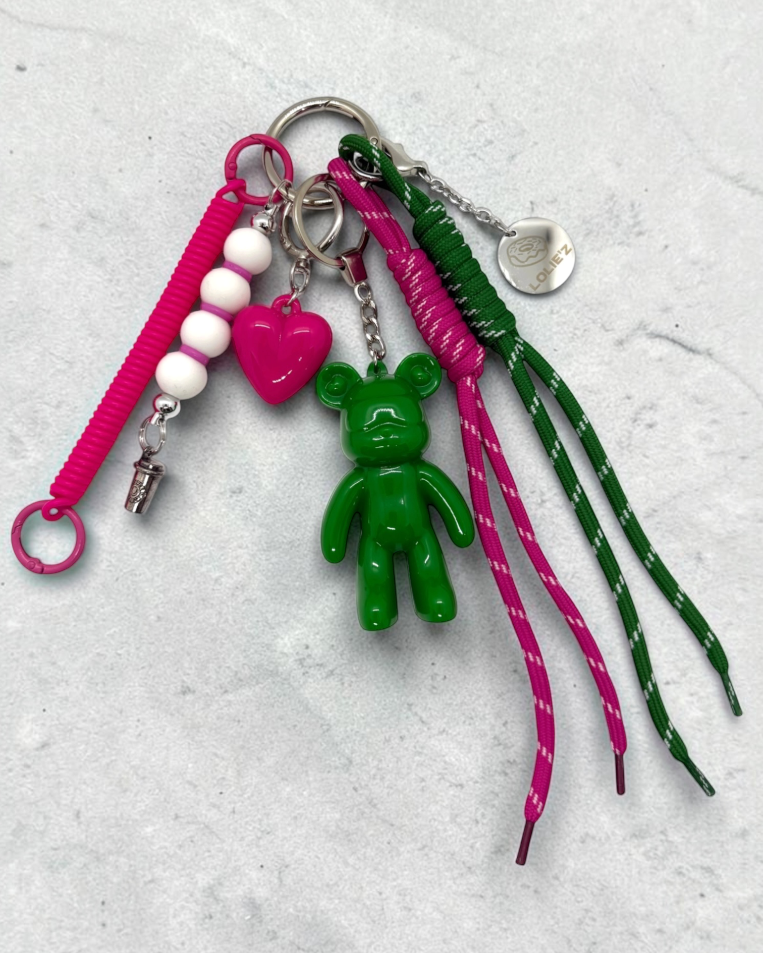 Grigri de sac Lolie’z vert et rose avec Teddy’z en résine époxy, cœur rose, cordons colorés, perles et médaille gravée, assemblé à la main en France.