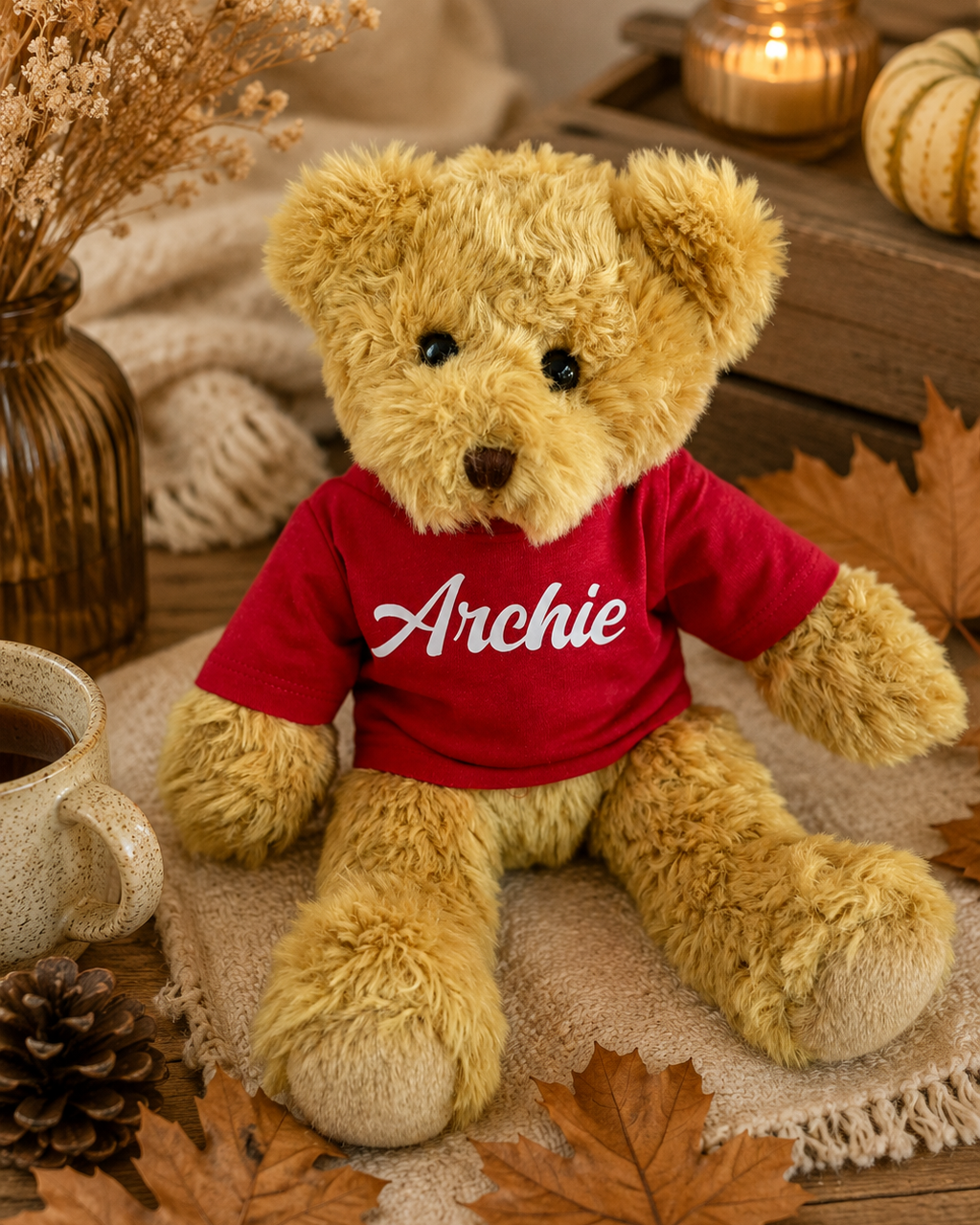 Peluche ours Archie avec t-shirt rouge personnalisable, mise en scène dans un décor d’automne chaleureux avec feuilles et matières naturelles.
